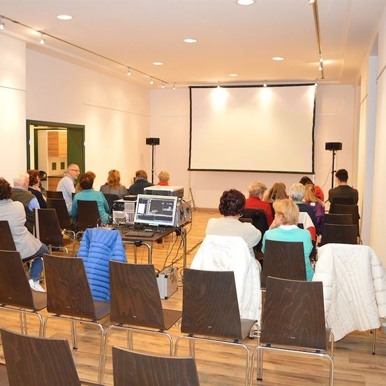 Eine Gruppe von Menschen sitzt in einem Raum mit einer Projektionsleinwand und Lautsprechern, wahrscheinlich bei einer Präsentation oder Besprechung.
