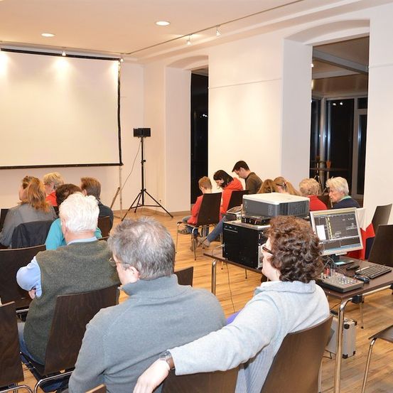 Eine Gruppe von Menschen sitzt in Stühlen in einem Raum mit einer Projektionsleinwand und einem Lautsprecher. Einige schauen auf einen Monitor.