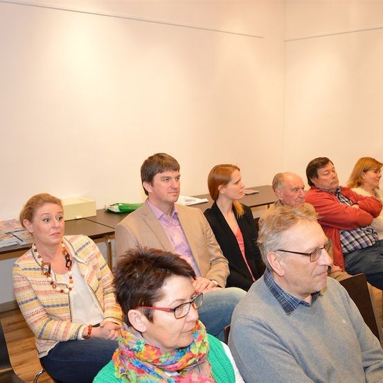 Eine Gruppe von Menschen sitzt in einem Raum, einige tragen Brillen und ein Mann steht.