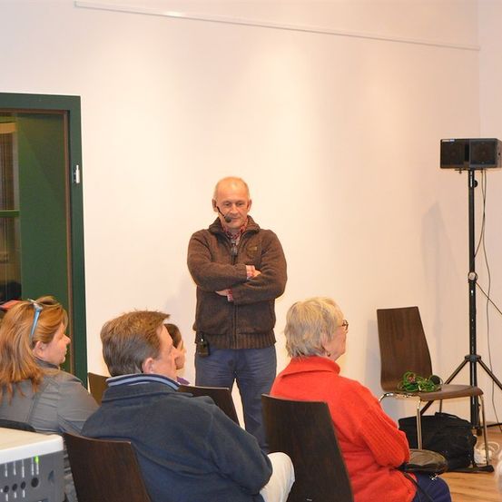 Ein Mann steht vor einer Gruppe von Menschen in einem Raum und trägt ein Mikrofon. Hinter ihm steht ein Lautsprecher auf einem Stativ. Das Publikum sitzt auf Stühlen und links im Raum befindet sich eine Tür.