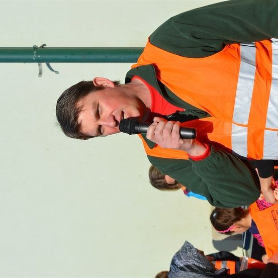 Ein Mann in einer orangefarbenen Sicherheitsweste spricht in ein Mikrofon, möglicherweise bei einer Veranstaltung.