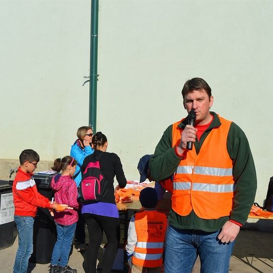 Ein Mann in einer Sicherheitsweste spricht in ein Mikrofon, während eine Gruppe von Kindern und Erwachsenen um einen Tisch mit verschiedenen Gegenständen steht.