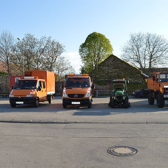 Vier Fahrzeuge, darunter zwei orangefarbene Nutzfahrzeuge, ein grüner Traktor und ein schwarzer Lkw, sind auf einem Parkplatz abgestellt. Bäume und ein Gebäude sind im Hintergrund zu sehen.
