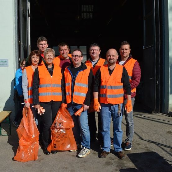 Eine Gruppe von Menschen in orangefarbenen Westen und Handschuhen steht vor einem Gebäude mit einem Schild, das 'CHAFTSHOP' besagt. Sie halten orangefarbene Taschen.