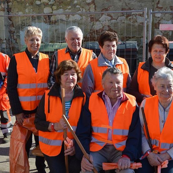 Eine Gruppe älterer Personen in orangefarbenen Sicherheitswesten und mit Stöcken, die vor einer Steinmauer und einem Metallzaun für ein Foto posieren.