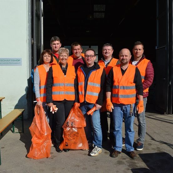 Eine Gruppe von Menschen in orangefarbenen Westen und Handschuhen steht vor einem Gebäude. Sie halten orange Plastiktüten. Hinter ihnen befindet sich ein Schild an der Wand.