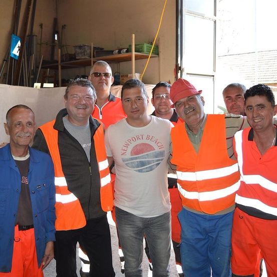 Eine Gruppe von Männern in Sicherheitswesten und Mützen posiert für ein Foto in einem Raum mit Regalen und einer Leiter im Hintergrund.