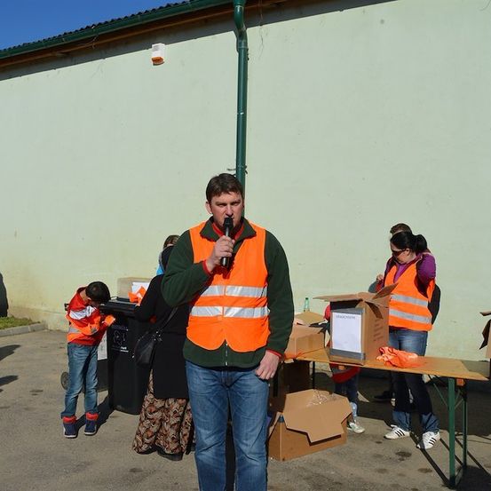 Ein Mann in einer orangefarbenen Warnweste steht vor einem Gebäude und hält ein Mikrofon. Hinter ihm sind mehrere Personen mit Aufgaben beschäftigt, darunter eine Frau, die durch Kisten sortiert.
