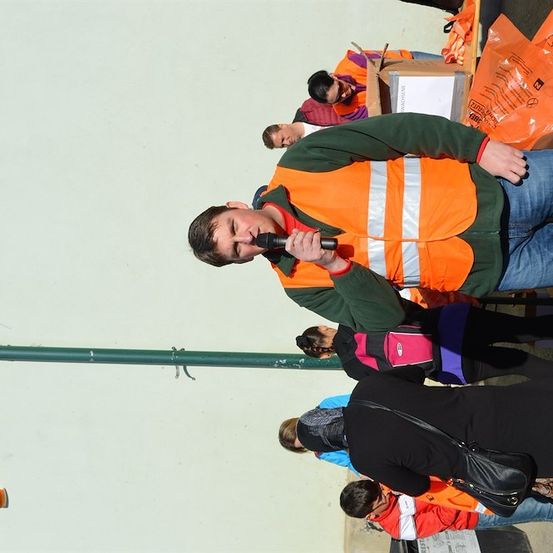 Ein Mann in einer orangefarbenen Warnweste spricht in ein Mikrofon, während mehrere Personen hinter ihm stehen. Der Ort scheint im Freien zu sein, mit einer weißen Wand im Hintergrund und verschiedenen orangefarbenen Plastiktüten.