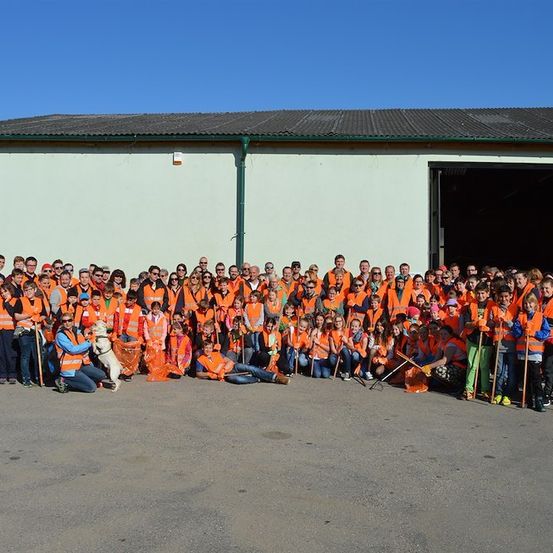 Eine große Gruppe von Menschen in orangefarbenen Westen und Sonnenbrillen posiert für ein Foto vor einem Gebäude. Einige halten Reinigungswerkzeuge und ein Hund ist anwesend.