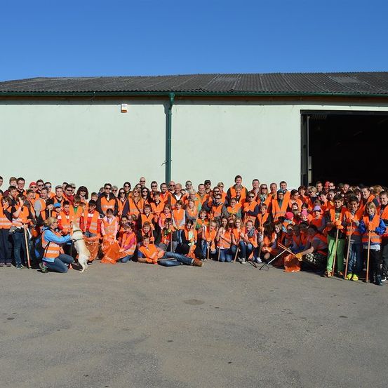 Eine Gruppe von Menschen in orangefarbenen Westen und Sonnenbrillen posiert für ein Foto vor einem Gebäude mit einer großen offenen Tür. Einige halten Stöcke und Taschen.
