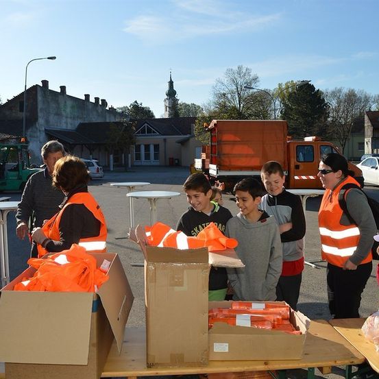 Eine Gruppe von Menschen in orangefarbenen Westen steht um einen Tisch mit Kisten voller orangefarbener Sicherheitsausrüstung. Im Hintergrund ist ein Gebäude mit einem Turm und ein Lastwagen zu sehen.