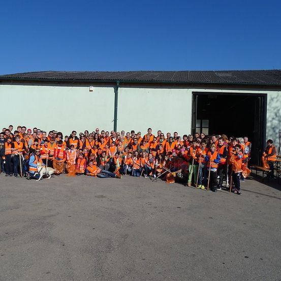 Eine große Gruppe von Menschen in orangenen Westen, mit Reinigungswerkzeugen, posiert für ein Foto vor einem Gebäude.