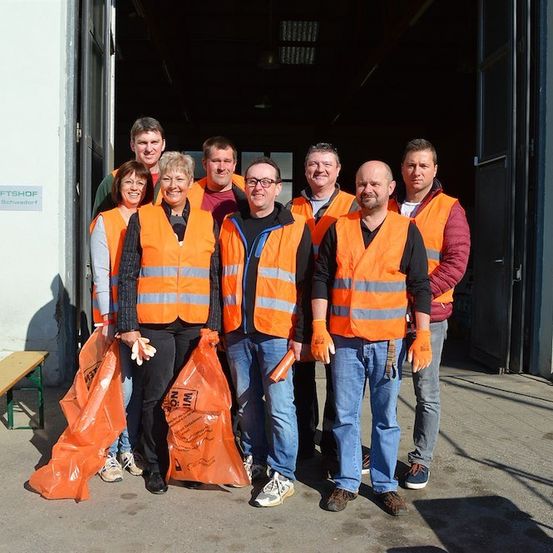 Eine Gruppe von Menschen in orangefarbenen Westen und Handschuhen steht vor einem Gebäude mit einem Schild, das 'ARTSCHAFSTSHOP' besagt. Sie halten orangefarbene Taschen und scheinen für ein Foto zu posieren.