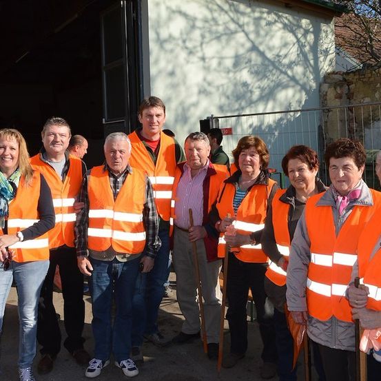 Eine Gruppe von Menschen in orangefarbenen Warnwesten steht vor einem Gebäude mit einem Metallzaun. Einige halten Stöcke und lächeln.