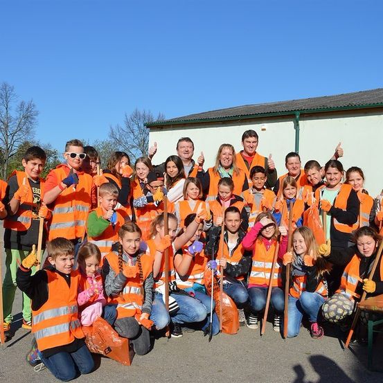 Eine Gruppe von Kindern und Erwachsenen in orangenen Sicherheitswesten posiert mit Reinigungswerkzeugen in einer sonnigen Außenumgebung.