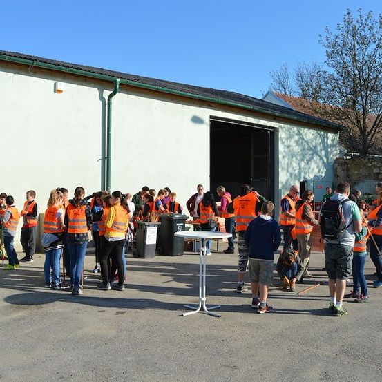 Eine Gruppe von Menschen in orangefarbenen Westen steht vor einem Gebäude mit einem Mülleimer, einem Tisch und einem Baum im Hintergrund.