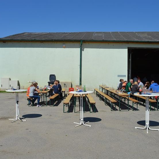 Eine Gruppe von Menschen sitzt an Tischen vor einem Gebäude mit offener Tür. Die Tische sind in Reihen angeordnet, und einige Leute stehen in der Nähe des Eingangs. Das Gebäude hat ein graues Dach und weiße Wände.