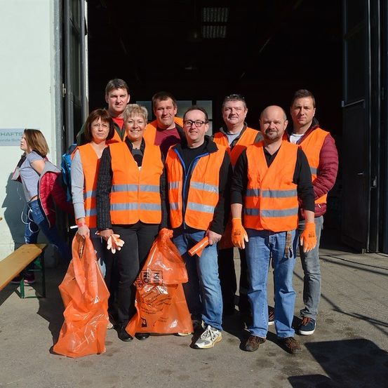 Eine Gruppe von Menschen in orangefarbenen Westen und mit orangefarbenen Tüten steht vor einem Gebäude. Einige lächeln, und eine Person hält eine Zigarette.