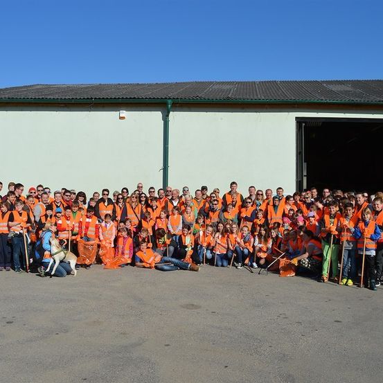 Eine große Gruppe von Menschen in orangefarbenen Westen und Sonnenbrillen posiert für ein Foto vor einem Gebäude. Einige halten Besen und Müllsäcke.