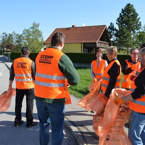 Eine Gruppe von Menschen in orangenen Westen steht am Straßenrand und hält orangene Beutel in der Hand. Sie nehmen an einer Müllsammelaktion teil.