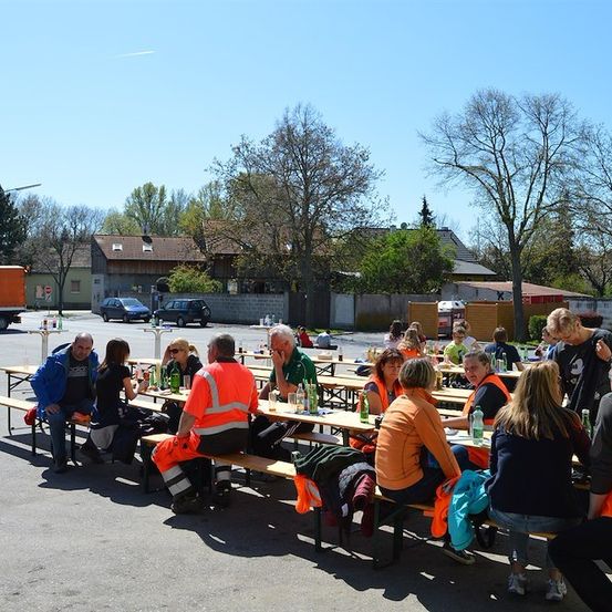 Eine Gruppe von Menschen in orangefarbenen Westen sitzt an Bänken auf einem Parkplatz mit Bäumen und Gebäuden im Hintergrund.