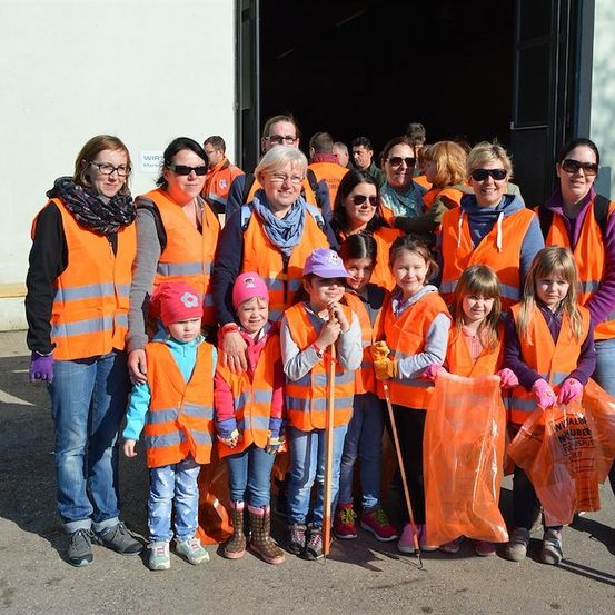 Eine Gruppe von Freiwilligen, einschließlich Kindern, die orange Westen tragen und Müllsäcke halten, stehen vor einem Gebäude. Sie lächeln und sehen aus, als wären sie bereit für eine Gemeinschaftsreinigungsaktion.