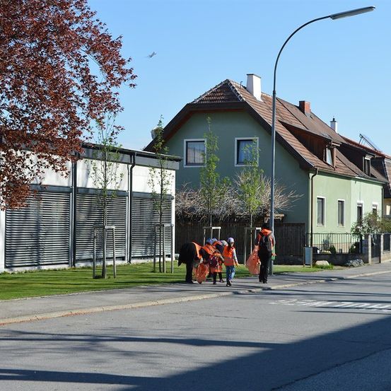 Eine Gruppe von Menschen in orangefarbenen Westen geht eine Straße entlang, mit einem Gebäude und Bäumen im Hintergrund.