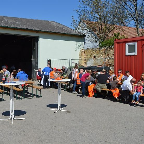 Eine Gruppe von Menschen sitzt um Tische in einem Außenbereich, möglicherweise bei einem Festival. Einige tragen orange Westen, und es gibt Bäume und Gebäude im Hintergrund.