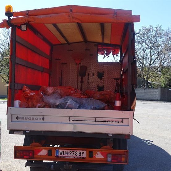 Die Rückseite eines Peugeot-Lkws mit offener Hecktür, die eine rote Abdeckung und verschiedene Gegenstände im Inneren zeigt, darunter Plastiktüten und ein Verkehrskegel.