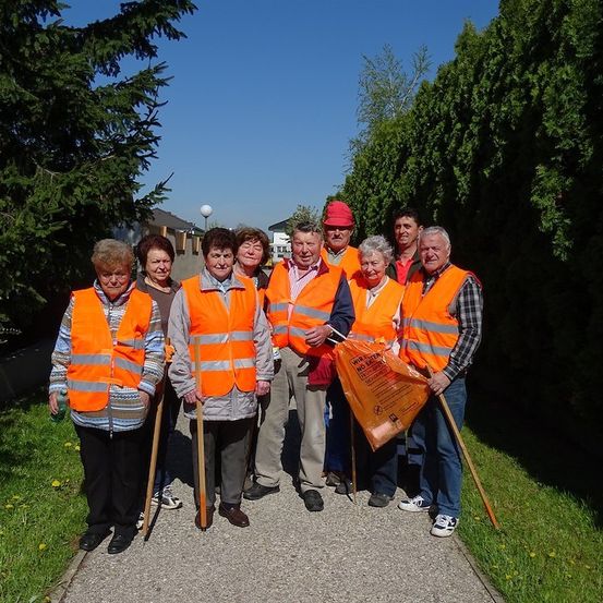 Eine Gruppe älterer Freiwilliger in orangefarbenen Westen und mit Stöcken, die auf einem Weg mit grünem Gras und Büschen im Hintergrund stehen.