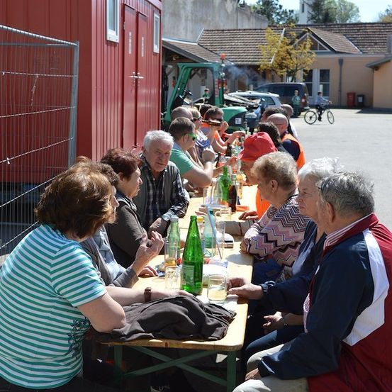 Eine Gruppe älterer Menschen sitzt draußen um einen Tisch herum, trinkt und plaudert. Sie sind vor einem roten Gebäude mit einem Metallzaun. Im Hintergrund sind geparkte Fahrzeuge und eine Person auf einem Fahrrad.