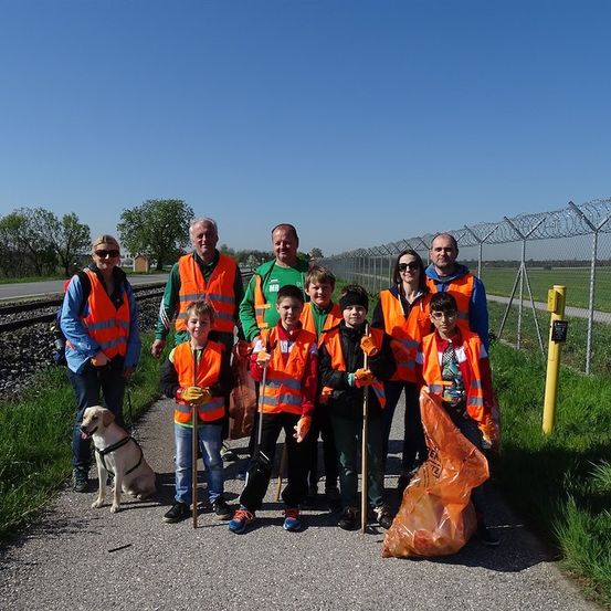 Eine Gruppe von Freiwilligen in orangefarbenen Westen und Handschuhen posiert für ein Foto, während sie Stöcke und Müllsäcke auf einem Kiesweg halten. Dahinter befindet sich ein Zaun mit Stacheldraht und ein Hund.