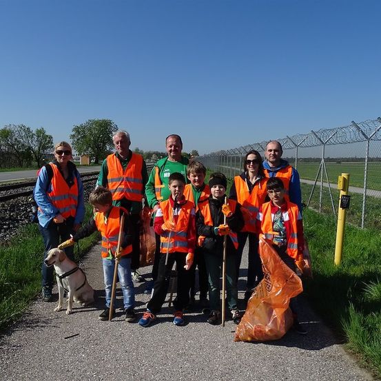 Eine Gruppe von Menschen in Sicherheitswesten und mit Stöcken steht auf einem Kiesweg. Sie nehmen wahrscheinlich an einer Gemeinschaftsaktion zur Müllsammlung teil. Ein Hund ist ebenfalls anwesend. Hinter ihnen befindet sich ein Zaun mit Stacheldraht, ein gelber Pfosten und ein Rasenbereich. Bäume und ein Gebäude sind in der Ferne zu sehen.