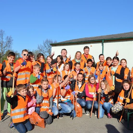 Eine Gruppe von Kindern und Erwachsenen in orangefarbenen Westen posiert für ein Foto vor einem Gebäude. Sie halten Stöcke in den Händen und einige tragen Handschuhe.