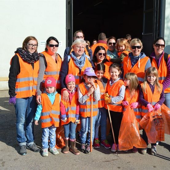 Eine Gruppe von Erwachsenen und Kindern in orangefarbenen Westen und Handschuhen steht vor einem Gebäude und hält Mülltüten.