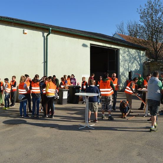 Eine Gruppe von Menschen in orangefarbenen Westen steht vor einem Gebäude, einige halten Stöcke und andere beugen sich. Ein Tisch und Mülleimer sind in der Nähe.