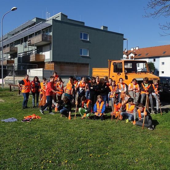 Eine Gruppe von Menschen in orangefarbenen Westen posiert für ein Foto auf einem Rasen mit einem LKW und Gebäuden im Hintergrund.