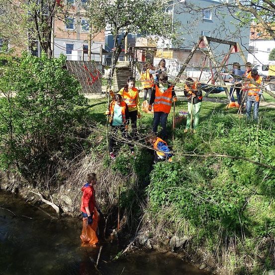 Eine Gruppe von Menschen in orangefarbenen Westen reinigt ein Flussufer, einige stehen am Ufer und andere im Wasser.