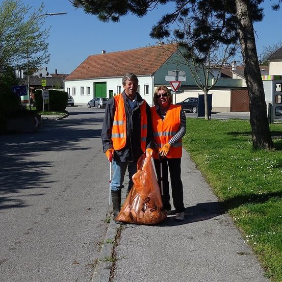 Ein Mann und eine Frau in orangenen Westen stehen auf einem Bürgersteig und halten einen Müllsack. Sie lächeln und scheinen für ein Foto zu posieren. Hinter ihnen befindet sich ein Rasenbereich mit einem Baum und einem Gebäude.