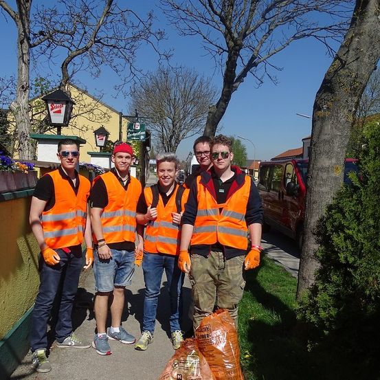 Fünf Männer in orange Weste und Handschuhen stehen auf einem Bürgersteig und lächeln für ein Foto. Dahinter ein Gebäude mit einem Schild und Bäumen. Ein Van und Müllsäcke sind in der Nähe.