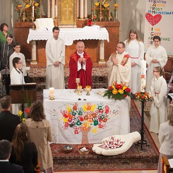 Eine religiöse Zeremonie mit Priestern und Altardienern in einer Kirche, umgeben von einer Gemeinde, mit einem geschmückten Altar mit Blumen und einem großen handbemalten Tuch.