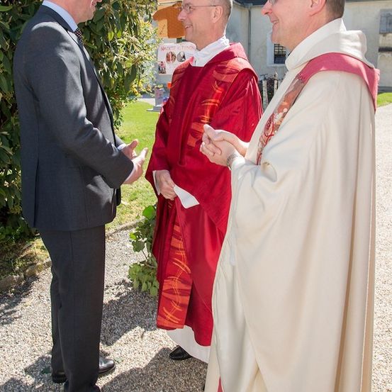 Drei Männer, einer im Anzug und zwei in geistlicher Kleidung, unterhalten sich vor einem Gebäude.
