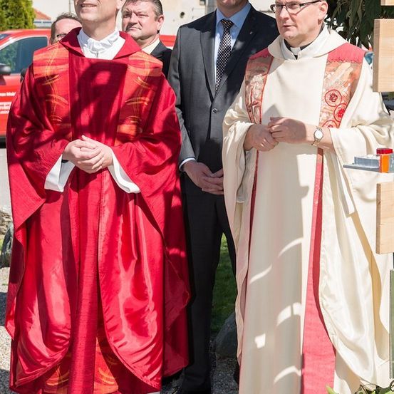 Zwei Priester in zeremonieller Kleidung stehen draußen, einer in Rot und der andere in Weiß, mit einem Mann in Anzug dahinter.