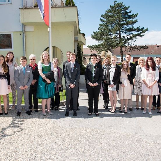 Eine Gruppe von Menschen in formeller Kleidung steht vor einem Gebäude mit einer Flagge und einem Baum im Hintergrund.