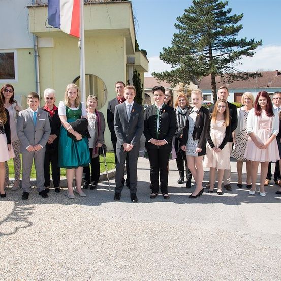 Eine Gruppe von Menschen in formeller Kleidung posiert für ein Foto vor einem Gebäude mit einer französischen Flagge an einem Fahnenmast.