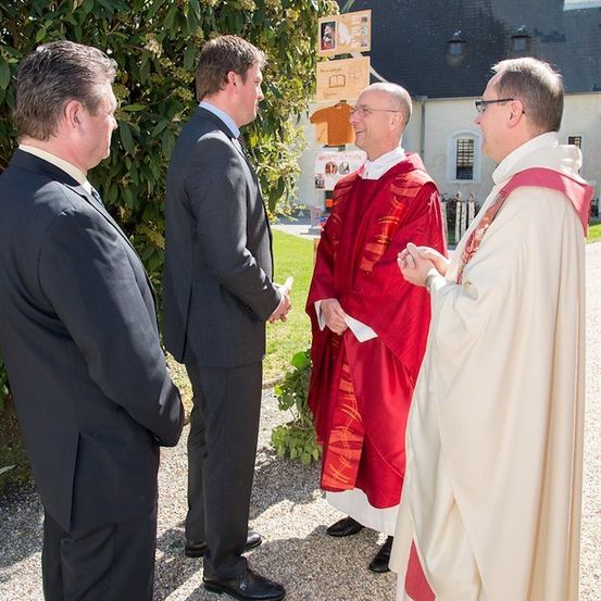 Drei Männer in Anzügen und zwei Priester in Gewändern unterhalten sich vor einer Kirche.