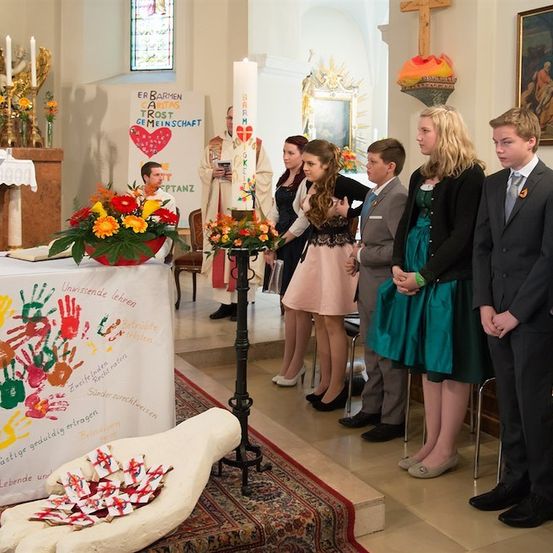 Eine Gruppe von Menschen in formeller Kleidung steht während einer Zeremonie in einer Kirche. Ein Priester steht am Altar, und ein Tisch mit Blumen ist in der Nähe. Eine große Handplastik steht auf dem Boden.