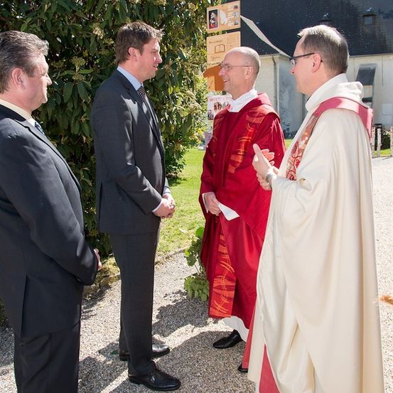 Vier Männer, zwei in Anzügen und zwei in geistlichen Gewändern, unterhalten sich vor einer Kirche.