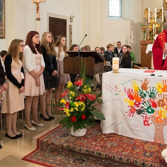 Ein Priester liest aus einem Buch, während eine Gruppe junger Frauen vor einem mit Blumen und Handabdrücken geschmückten Altar steht.